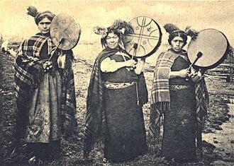 Three Machis standing side by side holding a Kultrun, which is a traditional Mapuche drum